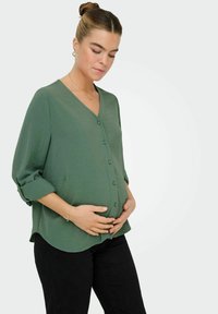 Blouse verte, légère, à boutons, avec un col en V, des manches retroussées et un tissu texturé, associée à un pantalon noir.