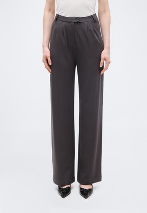 Femme portant un pantalon large taille haute gris foncé et des chaussures noires à bout pointu, debout devant un fond clair uni.