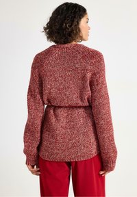 Kvindelig med krøllet hår iført en rød og hvid strikket sweater med et bælte, suppleret med røde bukser, vist fra bagenden mod en ensfarvet baggrund.