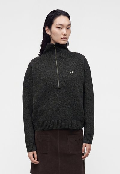 Femme portant un pull à col montant gris foncé avec une fermeture éclair devant et une jupe marron foncé avec des poches avant sur fond uni.