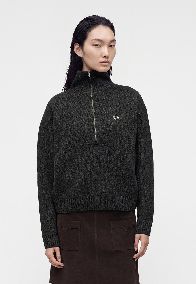 Femme portant un pull à col montant gris foncé avec une fermeture éclair devant et une jupe marron foncé avec des poches avant sur fond uni.