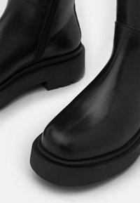 Botine din piele neagră cu o suprafață netedă, vârf unghiular și o talpă groasă, texturată, de culoare neagră. Disponibil cu un fermoar pe lateral pentru o purtare ușoară.