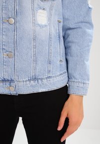 Jasnoniebieska kurtka dżinsowa z przetarciami noszona na czarnych spodniach, odsłaniająca środkową część ciała i luźną prawą rękę opartą na boku.