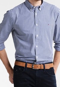 Camisa de cuadros azul y blanca con botones y cuello, combinada con un cinturón de cuero color beige. La camisa tiene un pequeño bolsillo en el pecho y mangas largas.
