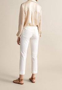 Jasnobeżowa satynowa koszula na guziki w połączeniu z białymi spodniami 7/8. Dopełnieniem stroju są brązowe sandały na paski. Widok od tyłu.