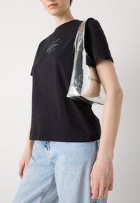 Zwarte katoenen t-shirt met geborduurd logo; lichtblauwe denim jeans; zilverkleurige metallic handtas met rits en mernaam erop gedrukt.