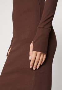 Brun ribbad långärmad klänning med figurnära passform, tummhål, och sömdetaljer, tillverkad av töjbar tyg.
