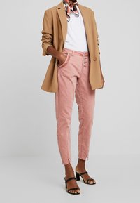 Ljusrosa avsmalnande byxor med sidfickor, med en knappslå framtill och en diskret slits vid vristerna. Bärs med en beige kavaj.