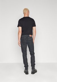 Blondýna v černém tričku, tmavých džínách a černých šněrovacích botách, stojící zády k fotoaparátu na jednoduchém světlém pozadí.