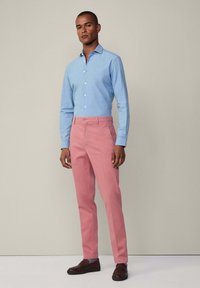 Hellblaues Hemd mit Knopfverschluss und Kragen, kombiniert mit rosa Hose und braunen Slippers. Die Hose hat eine glatte Stoffstruktur und eine figurnahe Passform.