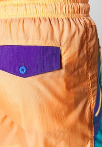 Barevné šortky z lehké oranžové tkaniny s texturovaným povrchem. Mají fialovou kapsu s modrým knoflíkem a kontrastními akcenty.