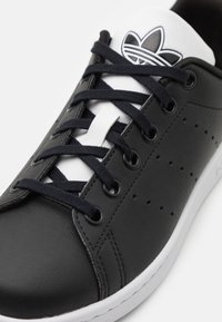 Zapatillas de cuero negro con acentos en blanco, puntera redonda, cordones planos negros, detalle perforado en el lado y un logo en blanco y negro en la lengüeta.