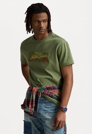 Oliven grøn bomulds t-shirt med rund halsudskæring og grafisk print. Kombinationen med slidte blå jeans og en flerfarvetternet skjorte, der er bundet om taljen.