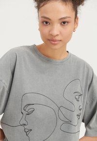 Ung kvinna med lockigt hår i en grå t-shirt med svart kontinuerlig linjekonst föreställande två abstrakta ansikten på framsidan.