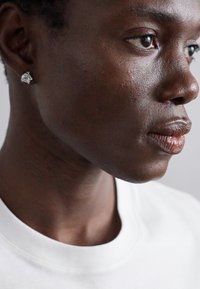 Pendiente de plata sobre una superficie de piel oscura y suave, con un primer plano de un rostro iluminado por luz natural y una camiseta blanca simple.