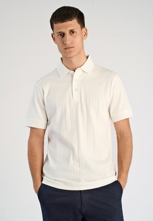 Crème kleurig polo-shirt met korte mouwen, voorzien van een textured verticale streepjespatroon en een drieknoopsluiting. Zwarte broek.