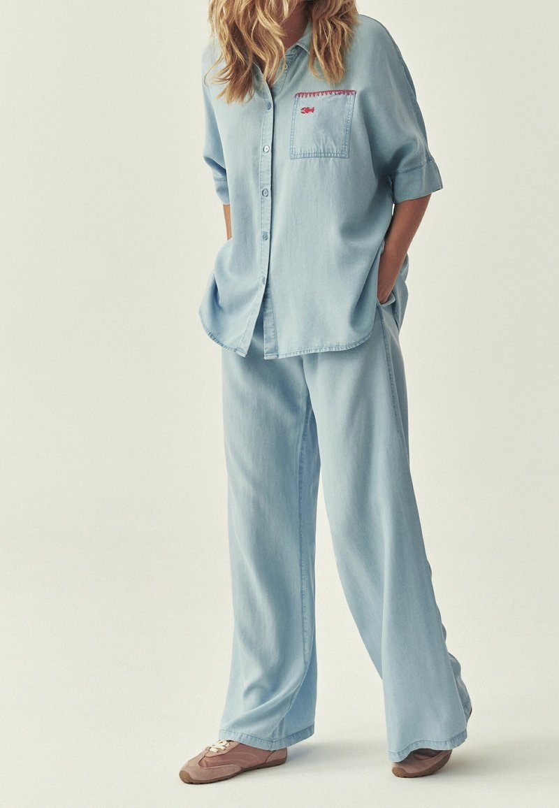 Personne portant une chemise ample bleu clair boutonnée et un pantalon à jambes larges, les mains dans les poches et des chaussures décontractées marron sur fond uni.