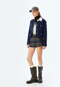 Giacca cropped navy con bottoni dorati, camicia bianca, minigonna a quadri, stivali al ginocchio marroni e berretto marrone. Tessuti texturizzati, look a strati.