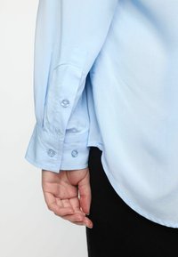 Camicia a maniche lunghe blu chiaro con polsini a bottone e un tessuto morbido e testurizzato. Presenta un orlo curvo e un sottile dettaglio di cucitura vicino al polsino.