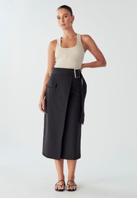 Ribana bež majica bez rukava u kombinaciji z crnom suknjom visokog struka s bočnom džepom i detaljima oko pojasa, glatke teksture i midi dužine.