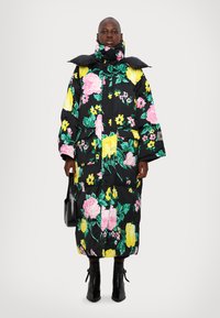 Abrigo acolchado con cuello alto; base negra adornada con grandes patrones florales en rosa y amarillo. Cuenta con dos bolsillos delanteros y botas negras.