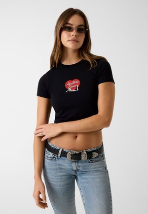 Młoda kobieta ubrana w czarny krótki T-shirt z czerwoną grafiką serca i napisem „Hysteric Queen”, niebieskie dżinsy, ozdobny czarny pasek oraz okulary przeciwsłoneczne.