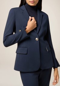 Blazer bleu marine à coupe ajustée, deux poches avant, fermeture à un bouton en ton doré, et une texture de tissu douce. Porté sur un col roulé noir.
