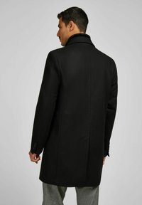 Cappotto di lana nera con colletto alto, vestibilità su misura e polsini abbottonati, caratterizzato da una texture liscia e una silhouette pulita.