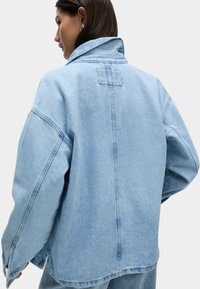 Veste en denim bleu clair à coupe décontractée, grand col et épaules tombantes. Présente une couture dans le dos simple et un design minimaliste.