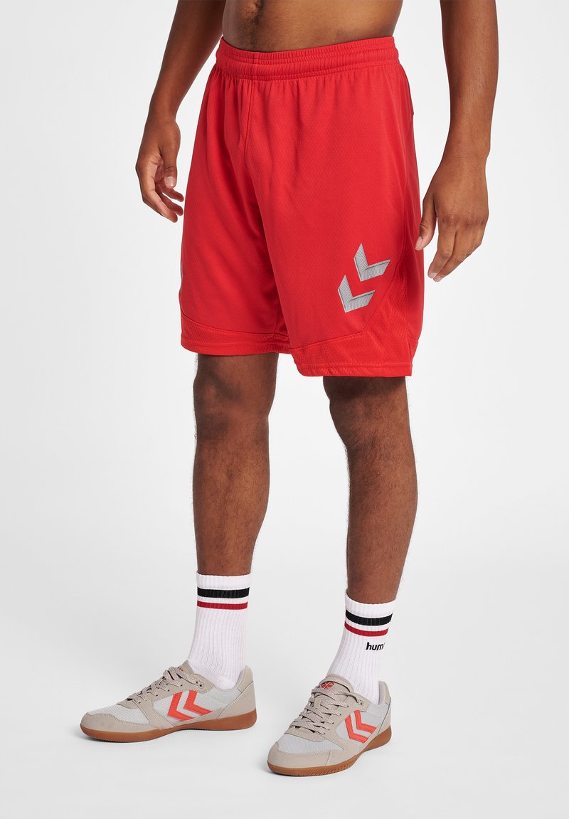 Röda idrottsshorts i ett strukturerat tyg, med grå chevrondetaljer. Modellen bär också grå skor och randiga strumpor.