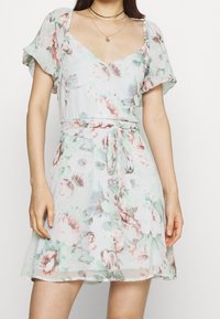 Robe fleurie en vert clair avec des motifs roses et blancs, dotée d'un décolleté en V, de manches flottantes et d'un détail de taille nouée. Tissu doux et léger.