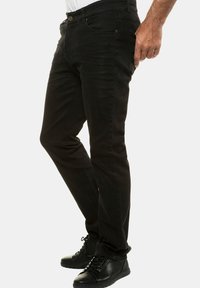 Jeans neri in denim con vestibilità slim, caratterizzati da cinque tasche e chiusura con bottone. Indossati con scarpe nere stringate, mostrati di lato.