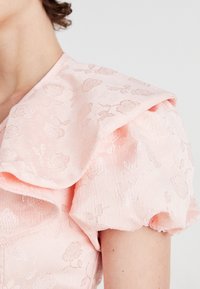 Lähikuva vaaleanpunaisesta kankaasta, jossa on kukallinen jacquard-painatus puffattulla lyhyellä hihalla ja suurella kerroksellisella kauluksella.