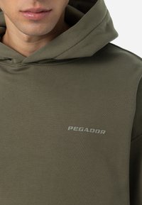 Olivengrøn hoodie lavet af bomuldsmateriale. Har en frontlomme og et gråt trykt logo "PEGADOR" på venstre brystområde. Glat tekstur.