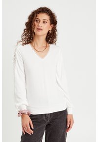 Top bianco a maniche lunghe con scollatura a V, dettagli in pizzo lungo le maniche e orlo a costine. Texture liscia, abbinato a jeans in denim scuro.
