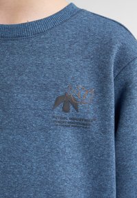 Sweat-shirt bleu en tissu texturé, avec un col côtelé. Comprend un logo imprimé en noir et orange sur la poitrine.