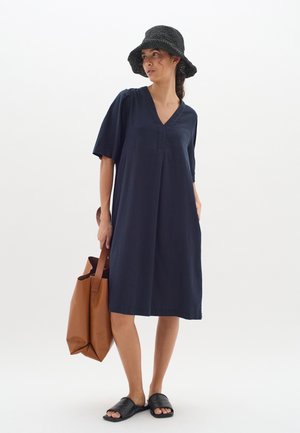 Rochie din in de culoare bleu marin, cu mâneci scurte și decolteu în V, având o croială lejeră. Asortată cu o geantă marron și sandale negre cu slip-on.