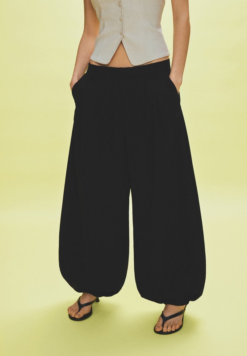 Personne portant un pantalon noir à jambes larges avec des ourlets pincés, un gilet beige sans manches boutonné et des sandales noires à bouts ouverts, debout sur un sol jaune pâle.