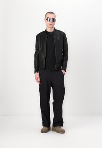 Giubbotto di pelle nera con cerniera, t-shirt nera, pantaloni cargo neri e stivali color sabbia. Gli accessori includono occhiali da sole argentati e orecchini.