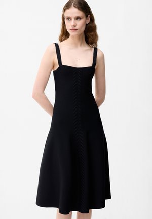 Kobieta w czarnej sukience midi bez rękawów z szerokimi ramiączkami i subtelnymi pionowymi teksturowanymi detalami na środku.