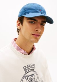 Ανοιχτό μπλε βαμβακερό καπέλο με καμπυλωτό γείσο, που φέρει ένα μικρό έμβλημα. Λευκή μπλούζα με στρογγυλή λαιμ όκοψη και κόκκινη ριγέ γιακά, με κεντημένη λεπτομέρεια.