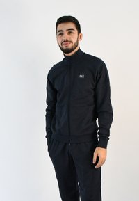 Sudadera de cremallera en color navy, hecha de una tela suave, con cuello tipo mock, puños acanalados y un logo bordado en el pecho. Combinada con pantalones a juego.