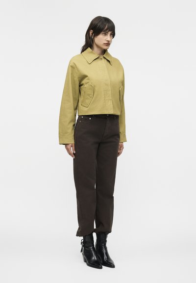 Γυναίκα που φοράει κοντό μπουφάν ελιάς, παντελόνι σκούρο καφέ με ψηλή μέση και μαύρα μποτάκια με τακούνι, στέκεται μπροστά σε απλό λευκό φόντο.