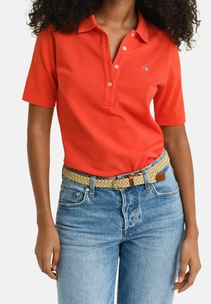 Kobieta ubrana w czerwoną koszulkę polo z krótkim rękawem, wpuszczoną w niebieskie dżinsy z plecionym beżowym paskiem, stojąca na jednolitym tle.