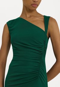 Grön ärmlös klänning med asymmetrisk halsringning, texturerad drapering på sidan, slät tyg och smal passform som framhäver kroppens form.
