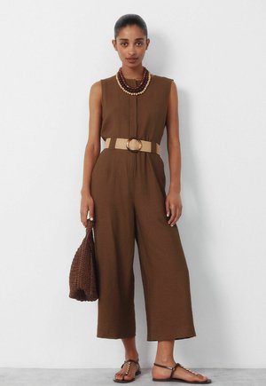 Vrouw draagt een mouwloze, bruine jumpsuit met brede pijpen, een beige riem, gelaagde kralenkettingen, houdt een gevlochten bruine tas vast en draagt donkere sandalen.