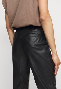 Svarta läderbyxor med en slät yta, med två bakfickor och en åtsittande midja. Modellen bär en brun topp.