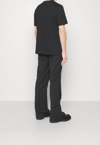 Černé bavlněné tričko s krátkými rukávy a kulatým výstřihem, kombinované s černými kalhotami s širokými nohavicemi a robustními černými botami, viděno ze zadu.