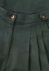 Donkergroene plooibroek met een tailleband met twee bruine knopen, zijzakken en een gestructureerde stof.