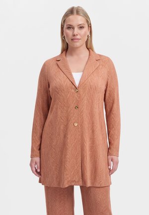 Frau trägt einen rostfarbenen, strukturierten langen Blazer mit drei goldenen Knöpfen und passenden Hosen vor einem einfarbigen hellen Hintergrund.